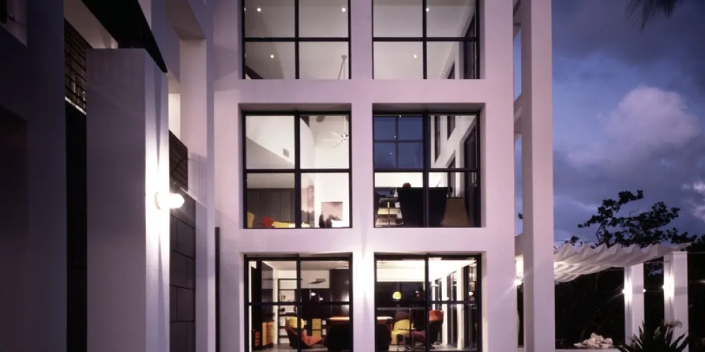A captivating evening view of the home's contemporary facade showcases expansive grid-like windows, warmly illuminating interior spaces. This design harmoniously connects the outdoor and indoor environments, creating an inviting ambiance.