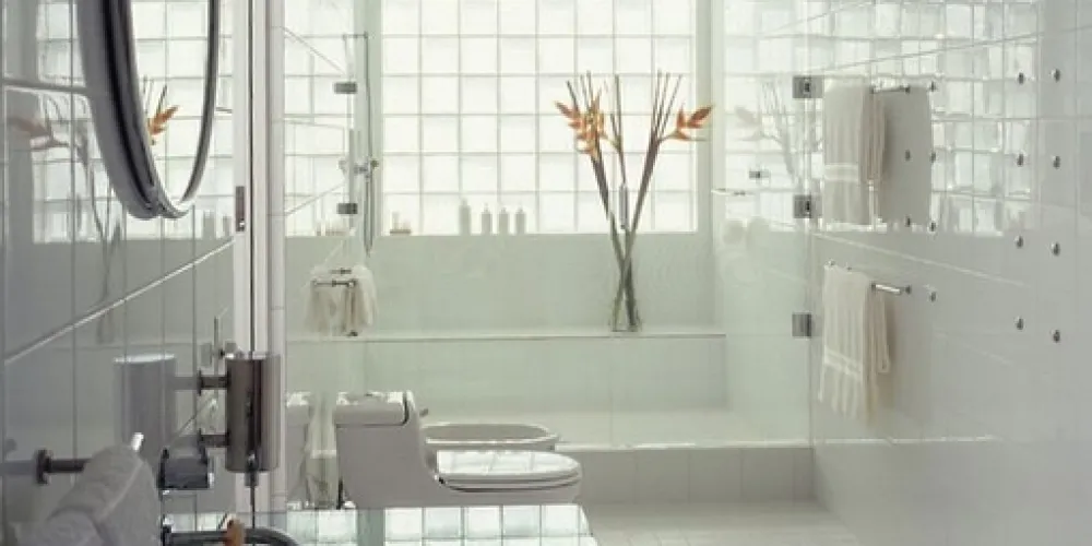 A pristine white bathroom with glossy tiles and glass accents creates a serene, modern space. Stainless steel fixtures and natural light enhance its clean, refreshing, and sophisticated appeal.

