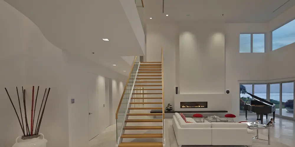 This living room is characterized by a minimalist, open-plan design with a striking modern staircase featuring glass railings and wooden steps. The double-height ceiling enhances the sense of space, while large windows let in natural light and offer stunning views.
