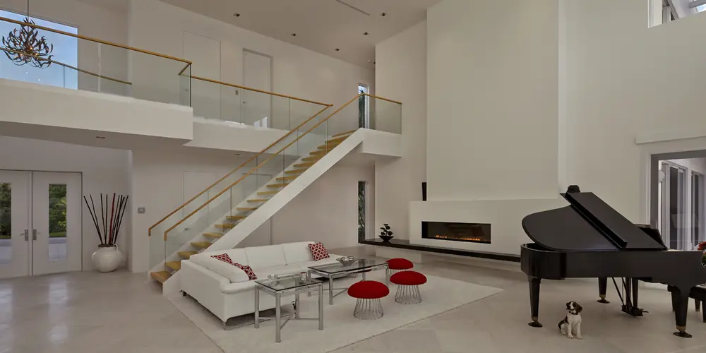 The living room offers a harmonious blend of minimalist design and sophistication, featuring a grand piano, decor, and ample open space. The double-height ceiling and expansive views continue the theme of elegance and openness.

