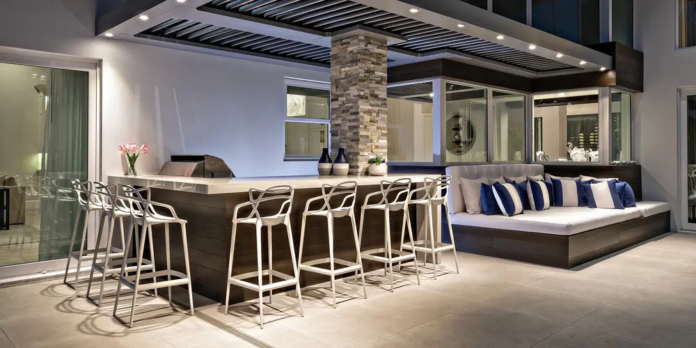 This outdoor bar area combines functionality with style, featuring a bar island under a pergola-like structure that provides shade and focus. Integrated seating with plush cushions enhances the entertaining space, ideal for hosting gatherings.