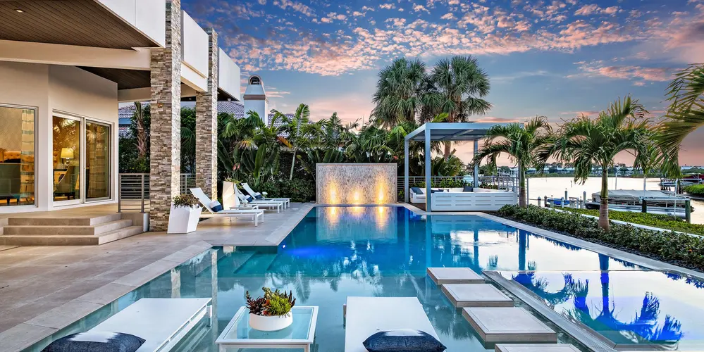 This poolside scene showcases a luxurious oasis designed for relaxation and entertainment. The integration of a jacuzzi with the main pool allows for seamless connectivity and flexibility in aquatic leisure. A boat dock visible in the background emphasizes the home's connection to the waterfront lifestyle, while a fountain feature adds a dynamic visual and auditory element to the landscape, enhancing the overall ambiance.