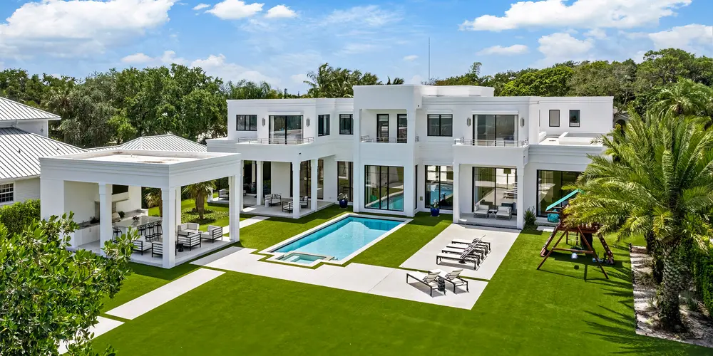 The backyard of this sophisticated residence showcases a seamless integration of luxury and functionality, centered around a pristine swimming pool that acts as the focal point of outdoor activity. The modern white facade of the house extends into the exterior space, offering a harmonious continuation of style. Spacious patios and meticulously manicured lawns provide ample space for relaxation and entertainment, surrounded by lush greenery that enhances privacy and adds to the tropical oasis vibe.