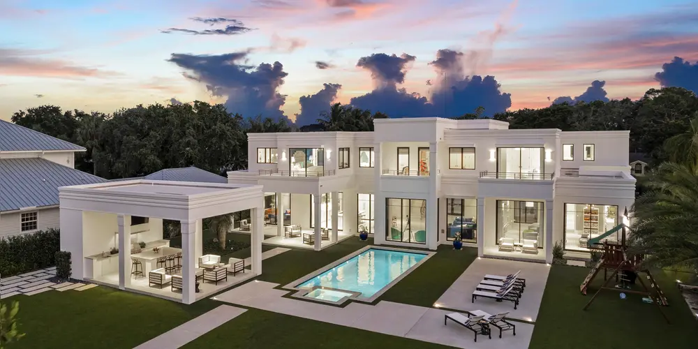 As twilight descends, the home's architecture is beautifully illuminated, highlighting its elegant features and creating an inviting ambiance. The strategic lighting enhances the structural lines and the pristine white facade, making the property stand out against the backdrop of a darkening sky. The pool reflects the subtle glow, adding a serene and luxurious aura to the overall setting. Together, these elements transform the home into a visually stunning nighttime retreat, offering a perfect blend of tranquility and sophistication.