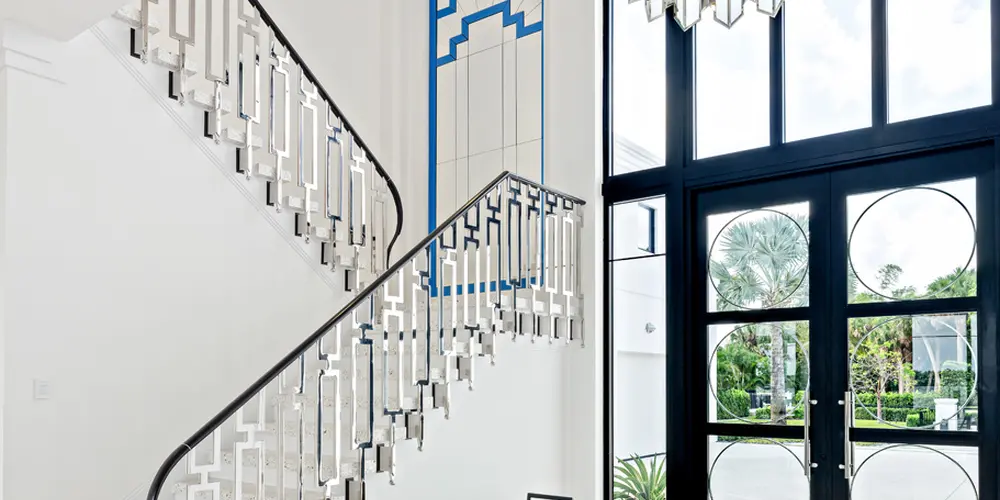 The interior of this building presents a sweeping sense of openness and luxury, characterized by high ceilings and spaciousness. The grand staircase, a focal point of the interior, is paired with a stylish banister and a contrasting geometric wall feature, creating a harmonious blend of traditional and modern aesthetics. Full-height windows allow natural light to flood the space, enhancing the white color palette and terrazzo flooring. A sleek, minimalist bench adds a touch of color and functionality to the foyer.