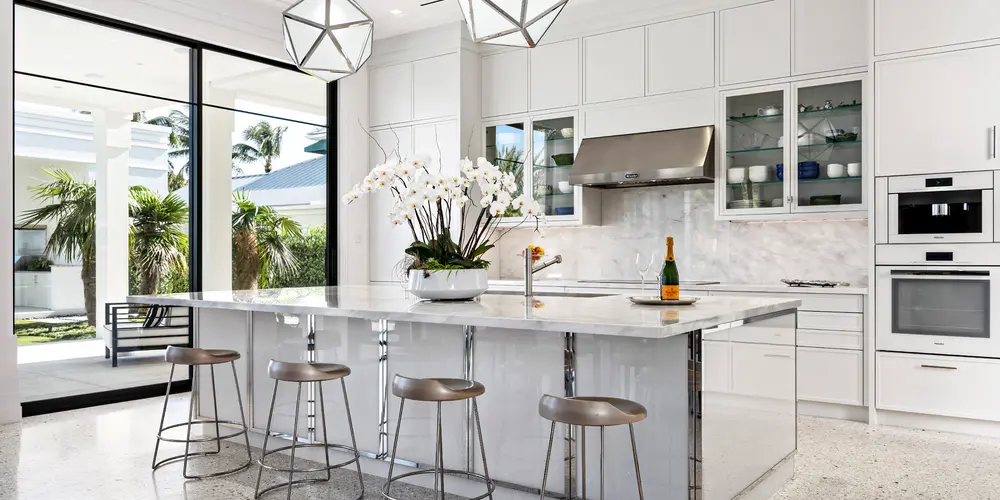 This open concept modern kitchen embodies elegance and functionality, serving as both a culinary hub and a social gathering space. Dominated by a sleek white palette, the kitchen's design emphasizes clean lines and a minimalist aesthetic while ensuring every function is met with style and precision. The large kitchen island doubles as a prep area and casual dining spot, inviting interactions and creating a sense of community within the home. Expansive glass doors blur the boundaries between indoor and outdoor living, enhancing the kitchen's spaciousness and its connection to the surrounding environment.