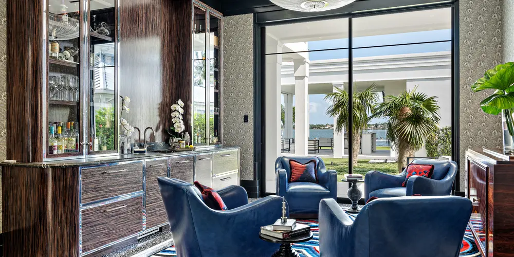 The luxurious lounge area exudes sophistication and comfort, offering a perfect blend of contemporary design and relaxing ambiance. The room is thoughtfully arranged with plush, deep blue armchairs encircling a chic central table, fostering an intimate and conversational atmosphere. Rich wood tones and intricate patterns on the cabinetry contrast elegantly with the sleek marble flooring, while large glass sliding doors provide a seamless transition to the outdoor spaces, ensuring the room is bathed in natural light and connected to the lush surroundings.