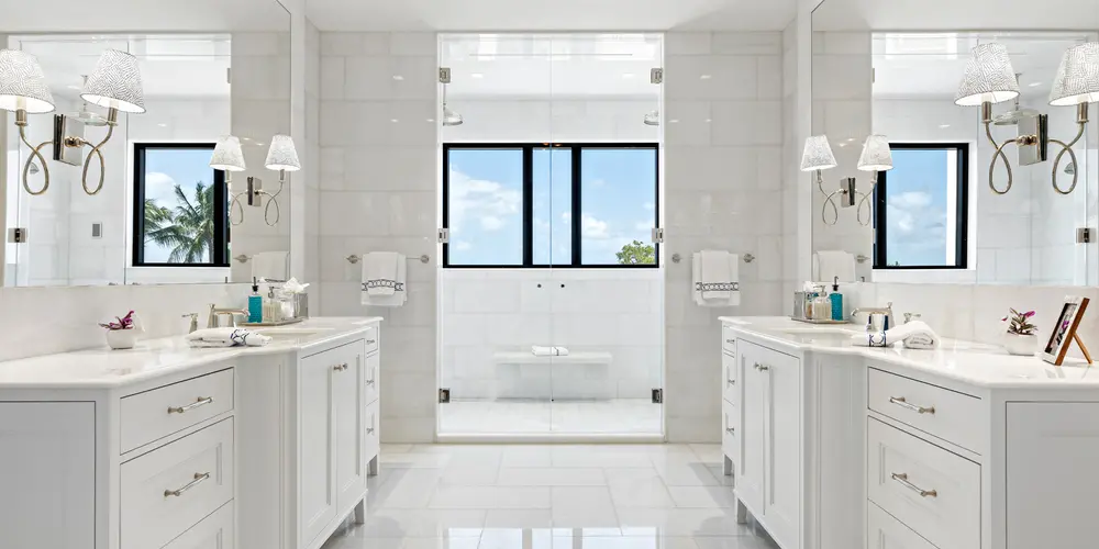 This master bathroom epitomizes luxury and functionality, offering a serene retreat within the home. Designed with symmetry and elegance in mind, the space features two white marble-topped vanities positioned opposite each other, creating a balanced and harmonious layout. A spacious glass-enclosed shower, centrally placed, acts as a focal point, framed by large windows that invite natural light and offer glimpses of the outdoors. The use of high-end materials and timeless design elements makes this bathroom a sanctuary of relaxation and refinement.