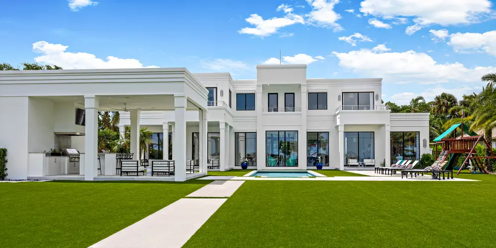 The rear exterior of this modern residence displays a seamless blend of architectural elegance and expansive outdoor living. The design features clean lines and a white facade, extending the interior’s contemporary aesthetic to the exterior. The open-air structure to the side offers a dedicated outdoor kitchen and lounge area, encouraging al fresco dining and relaxation. At the center, a sleek swimming pool provides a focal point for entertainment and leisure, surrounded by lush green lawns that contribute to a serene and open environment. The overall design emphasizes a seamless integration of indoor and outdoor spaces.