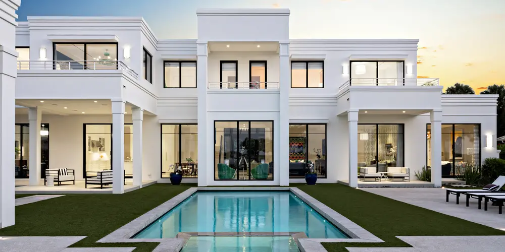 The rear facade of this modern residence, captured at sunset, showcases an exquisite blend of architectural precision and serene ambiance. The design is characterized by its symmetry and clean lines, with a prominent central swimming pool that draws attention and adds a touch of luxury. As the day transitions to evening, the illuminated interiors and outdoor lighting create a warm and inviting atmosphere, highlighting the home's elegance and attention to detail. The combination of white facades, expansive glass, and thoughtful landscaping enhances the sense of openness and tranquility.