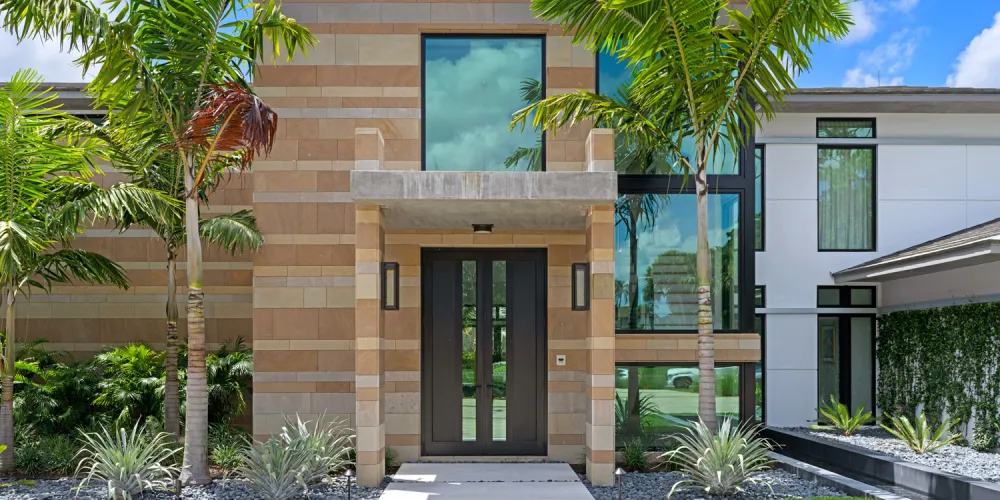 The front façade features a blend of modern materials with a clean, uncluttered design. The entryway is framed by tall palm trees, enhancing the home’s inviting yet sophisticated entrance.