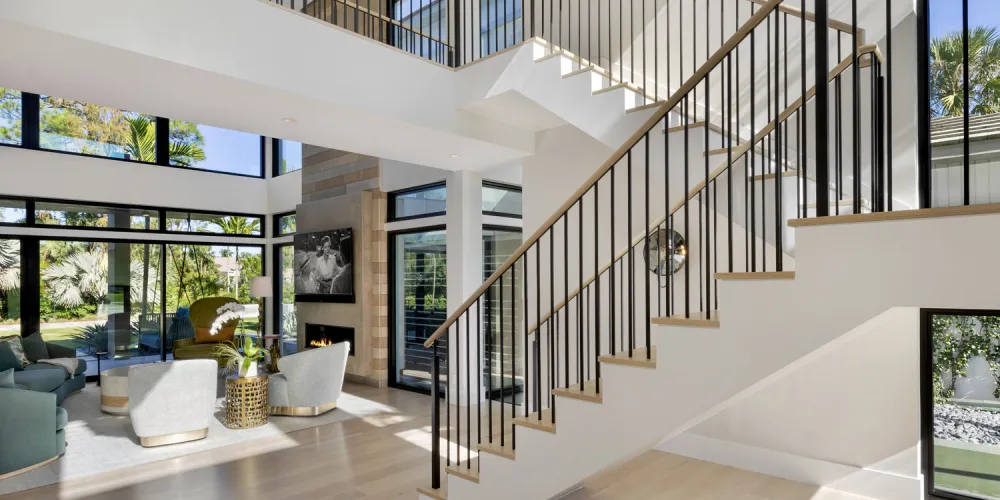 A beautifully designed staircase with sleek railing leads to a mezzanine level, emphasizing the home's modern and airy interior. Natural light streams through large windows, illuminating the space and highlighting the clean, minimalistic lines.