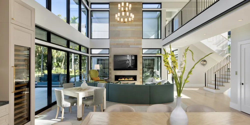 This expansive open-plan space merges the living and dining areas, bathed in natural light from the floor-to-ceiling windows. A modern chandelier accentuates the double-height ceiling, and light-toned furnishings complement the serene ambiance.
