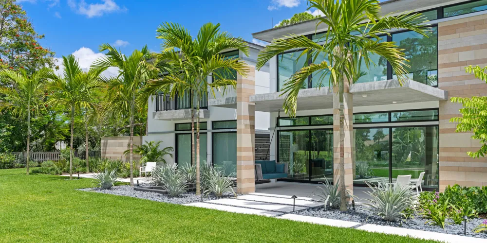 The exterior features a modern architectural style characterized by clean lines and expansive glass walls, complemented by stone accents. Tropical landscaping with palm trees and manicured lawns enhances the home’s natural appeal and integrates it with the lush surroundings.