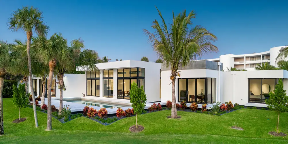 The house's exterior is marked by its striking white façade and grand, clear glass windows that reflect the vibrant blue sky. The surrounding tropical landscape, featuring well-maintained palm trees and shrubbery, contributes to an overall sense of tranquility and luxury.
