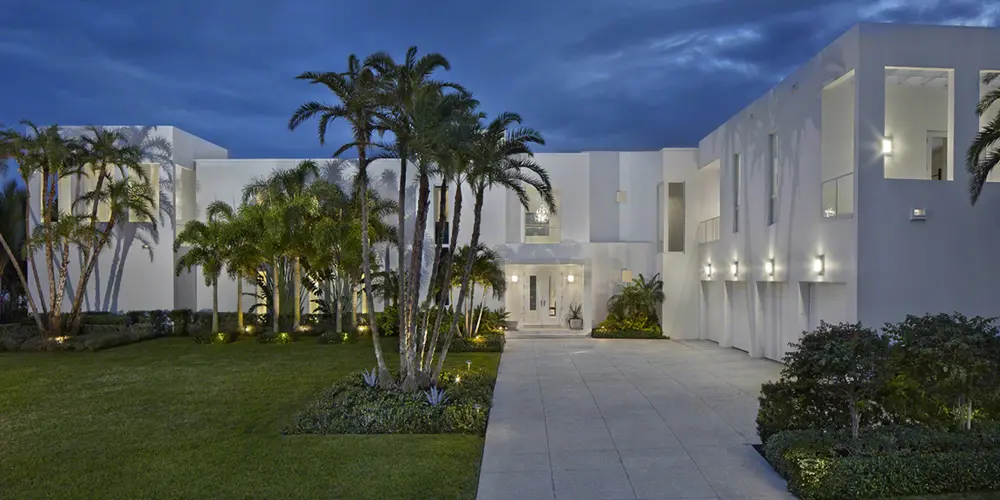  The entrance is defined by a striking facade with clean, white, geometric planes accented by shadows that play across its surfaces, giving depth and interest. The design incorporates vertical window slits that add a modern rhythm and welcome natural light. The landscaped path is lined with palm trees, leading to a sophisticated, grand entrance that seamlessly combines form with function.
