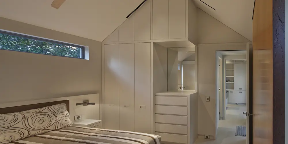 This bedroom utilizes vertical space effectively with a vaulted ceiling and integrated storage solutions. A sleek, minimalist design is maintained through neutral tones and clean lines.