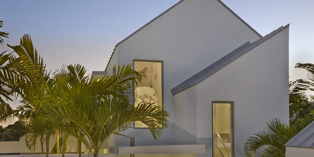 The exterior showcases modern design with sharp rooflines and tall windows. Lush palm trees are strategically placed, enhancing the architectural silhouette and providing a harmonious connection with nature.
