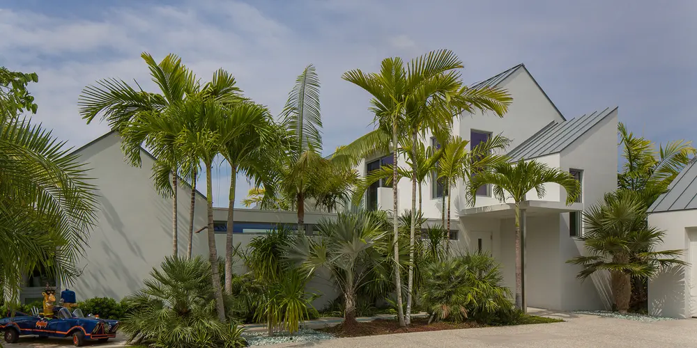 The daylight image captures the front facade’s integration with its lush environment, featuring dynamic rooflines and expertly arranged palm trees, enhancing the residence's stately presence.
