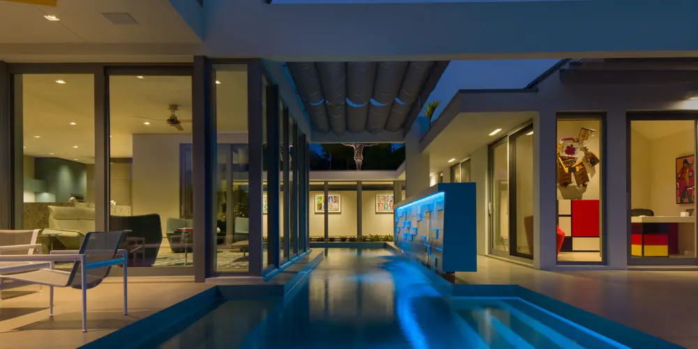 This perspective showcases a pool illuminated in vibrant lights, framed by large sliding glass doors, offering a seamless transition between indoor spaces and the illuminated outdoor retreat.