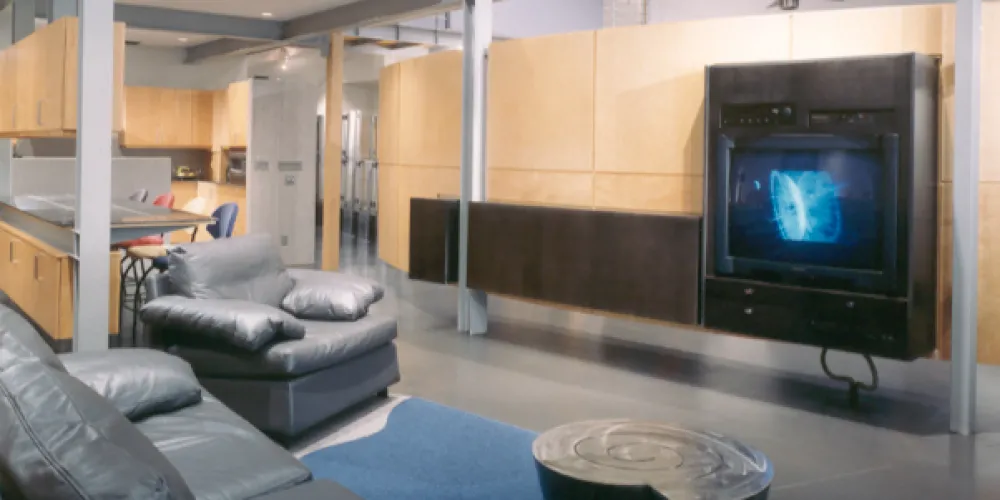 The living area is highlighted by the integration of technology, with a sleek entertainment center set against minimalist furnishings. Leather seating offers a comfortable, refined aesthetic. High ceilings and fans enhance the room's spaciousness, complemented by neutral tones for a seamless design flow.