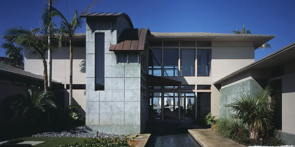 The entrance features a structured approach framed by modern architectural lines and a reflecting pool that gracefully integrates with the landscape. Natural foliage enhances this contemporary design, contributing to a welcoming and stylish front facade.
