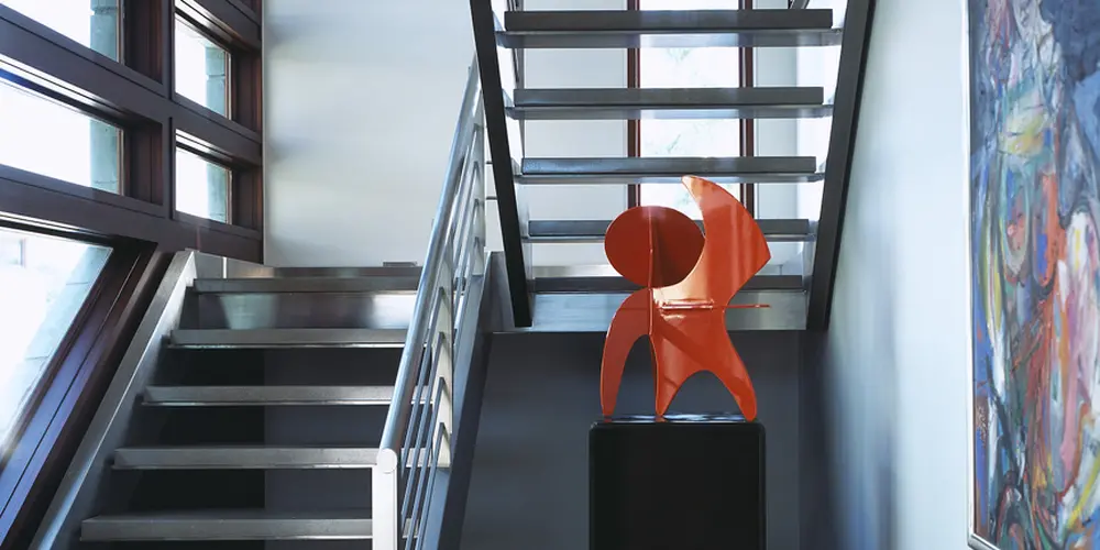 The staircase combines industrial materials with artistic expression, featuring open risers and a metal railing against a backdrop of modern art and bold color accents. The use of steel and concrete is softened by the strategically placed artwork and chic lighting, creating a dynamic, inviting environment.